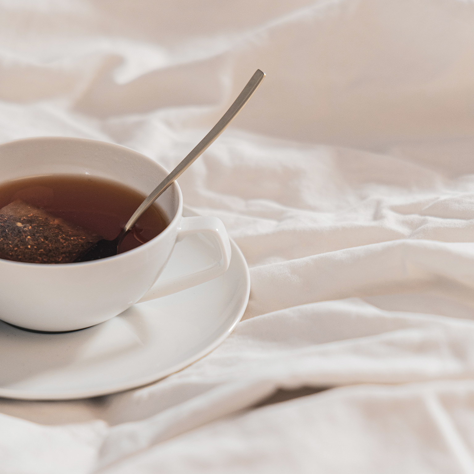 Side close-up of TAC White teacup with saucer, spoon on white sheet.